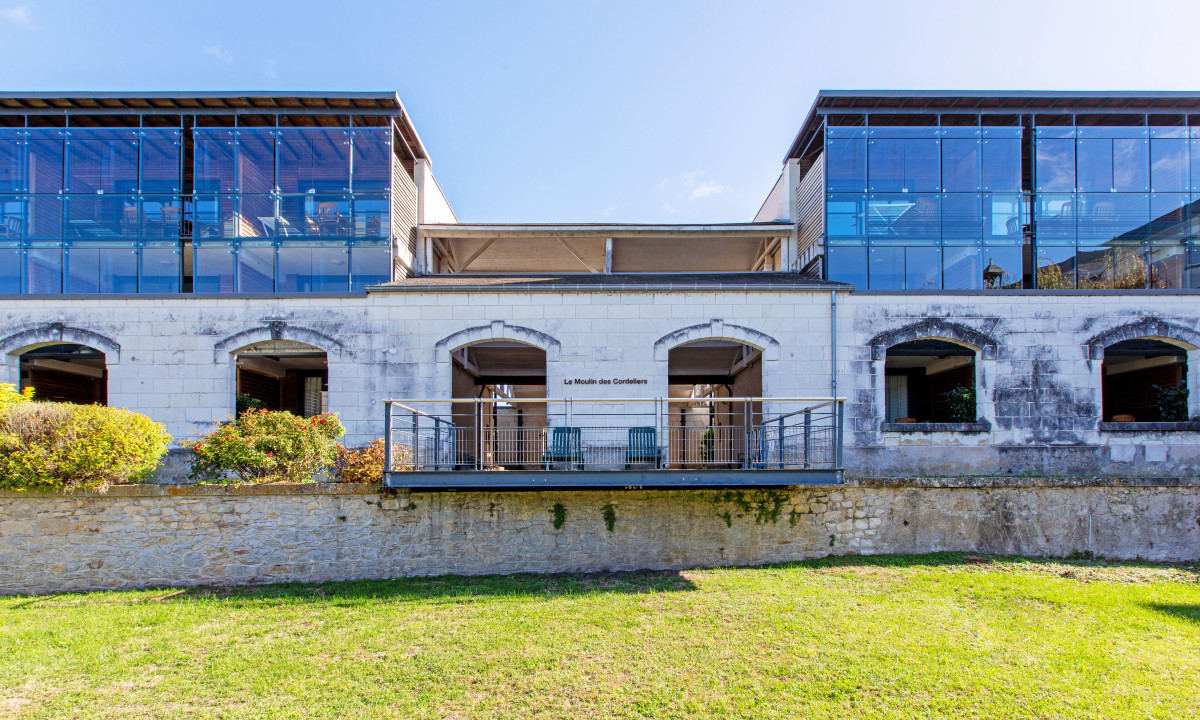 Stone building with glass windows and balconies, nestled in a garden on a sunny day at Le Moulin des Cordeliers