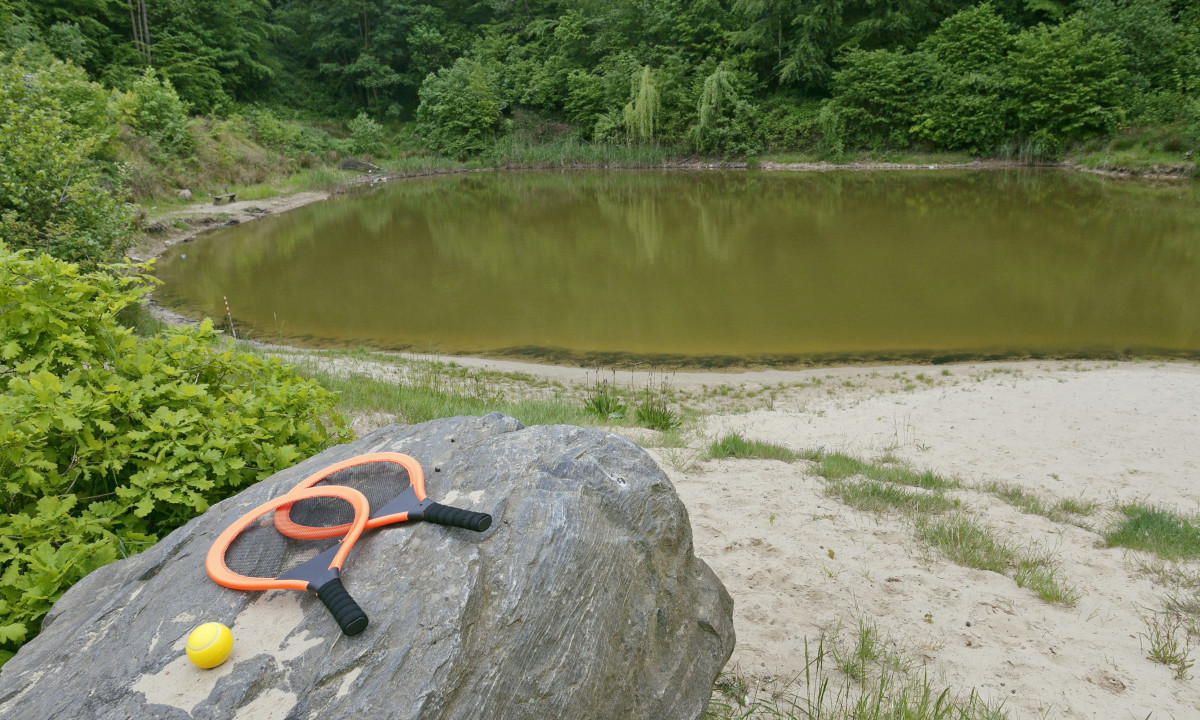At KNAUS Campingpark Wingst, orange badminton rackets and a yellow ball rest on a large stone next to a small lake encircled by trees.