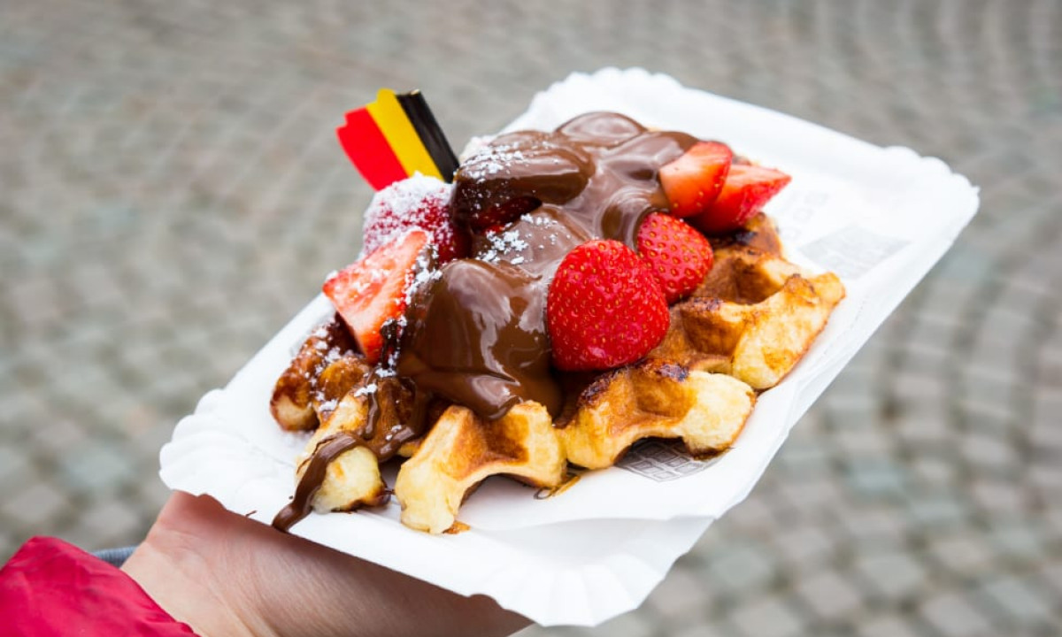 At Urban Gardens Gent, a waffle adorned with strawberries and chocolate is held in a hand above a cobblestone surface.