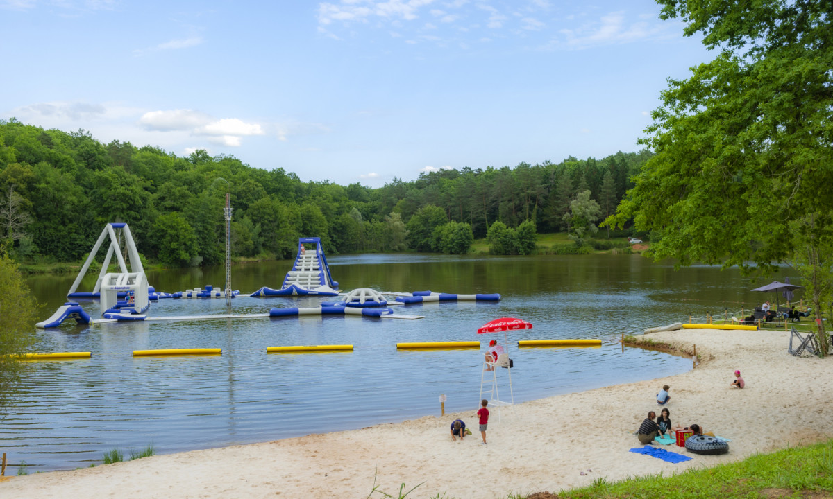 At Coucou, an inflatable water park drifts on a lake, encircled by a sandy shoreline and a verdant forested region where individuals are unwinding.