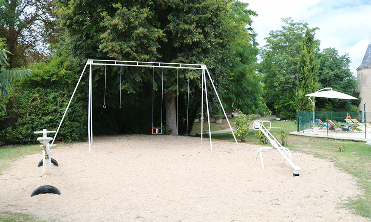 At Le Petit Trianon, playground apparatus remains idle in a sandy space encircled by verdant trees, adjacent to poolside reclining chairs.