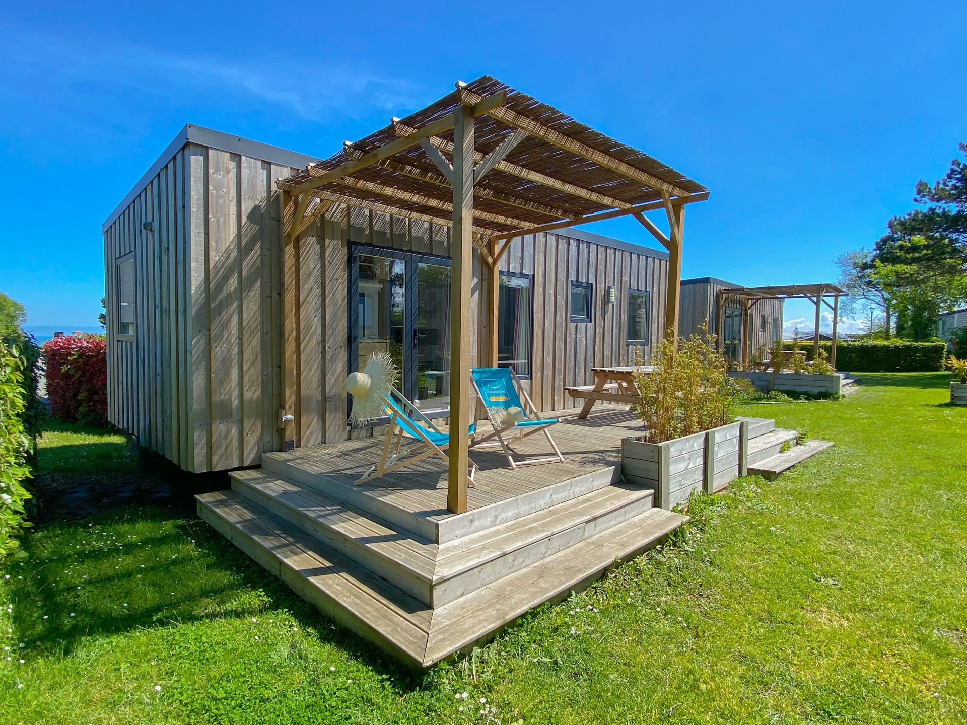 Wooden cabin with deck chairs and pergola, surrounded by greenery and adjacent cabins in a campsite setting at Seasonova Vittel