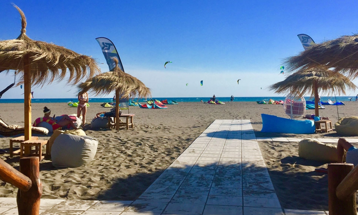 People lounging under straw umbrellas on a sandy beach with kitesurfers in the background at Safari Beach