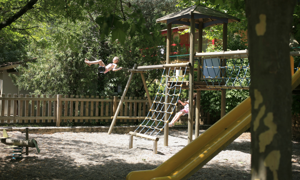 At La Clémentine, kids swing and climb on a play structure encircled by trees and a wooden barrier.