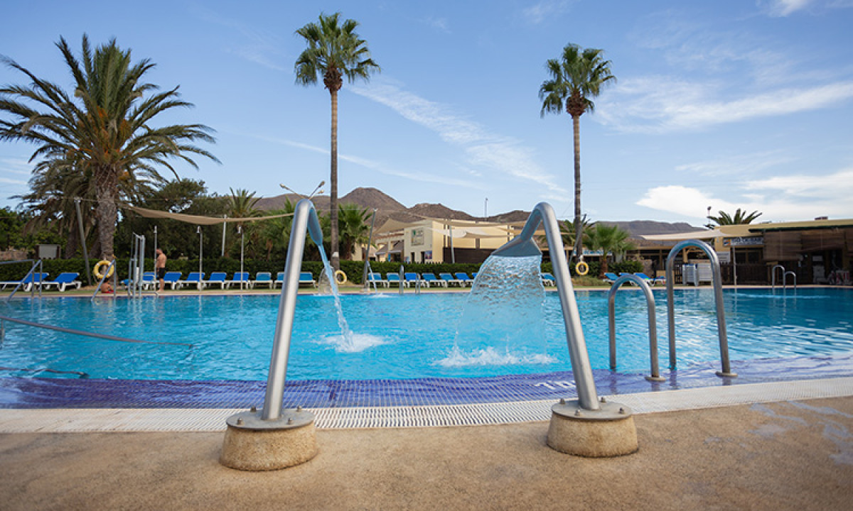At Camping Los Escullos, a pool with water jets streams, encircled by palm trees and mountains; deck chairs are arranged along the patio.