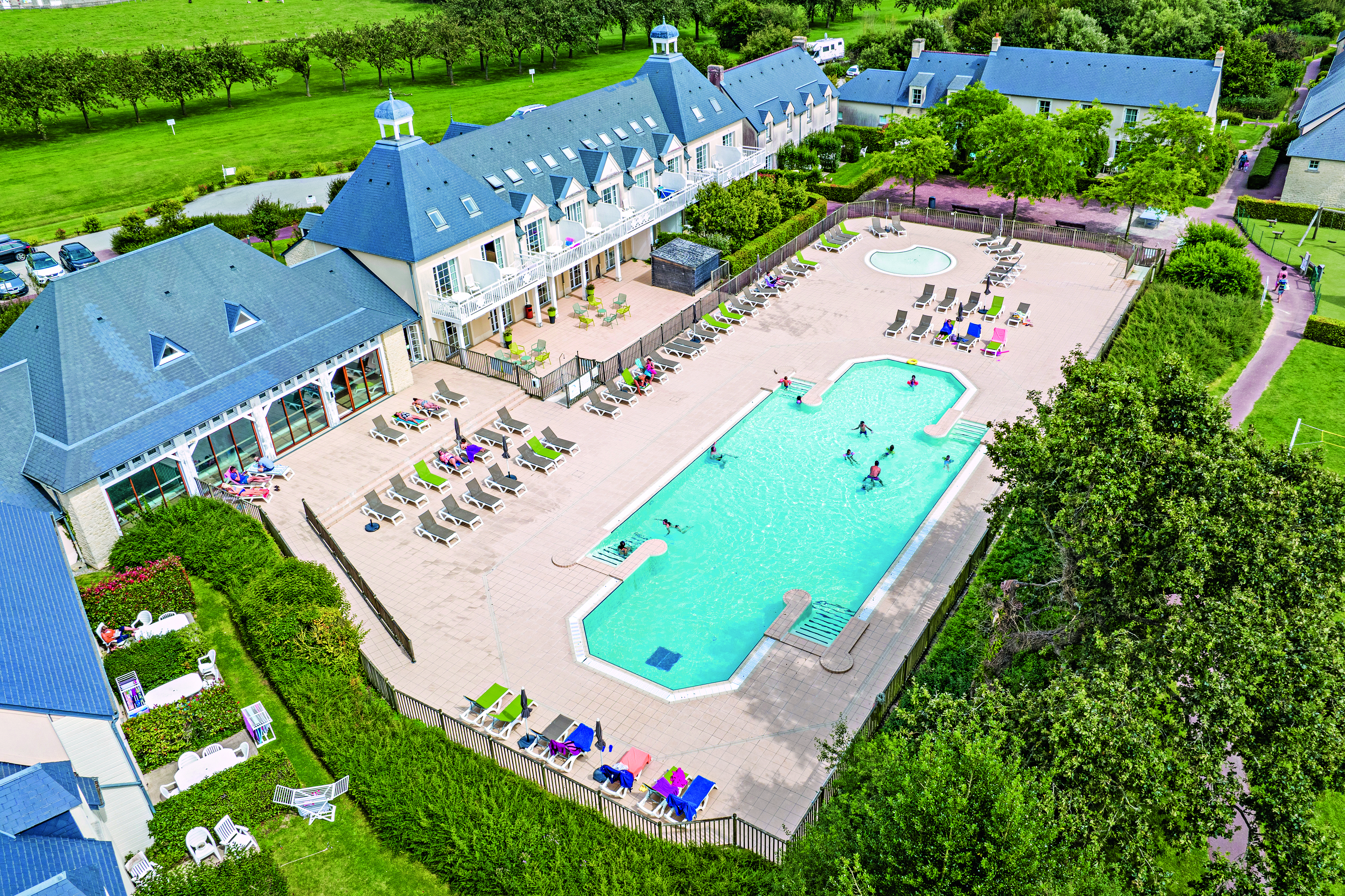 Swimming pool with sunbathers and swimmers beside a large building surrounded by green fields at Le Green Beach