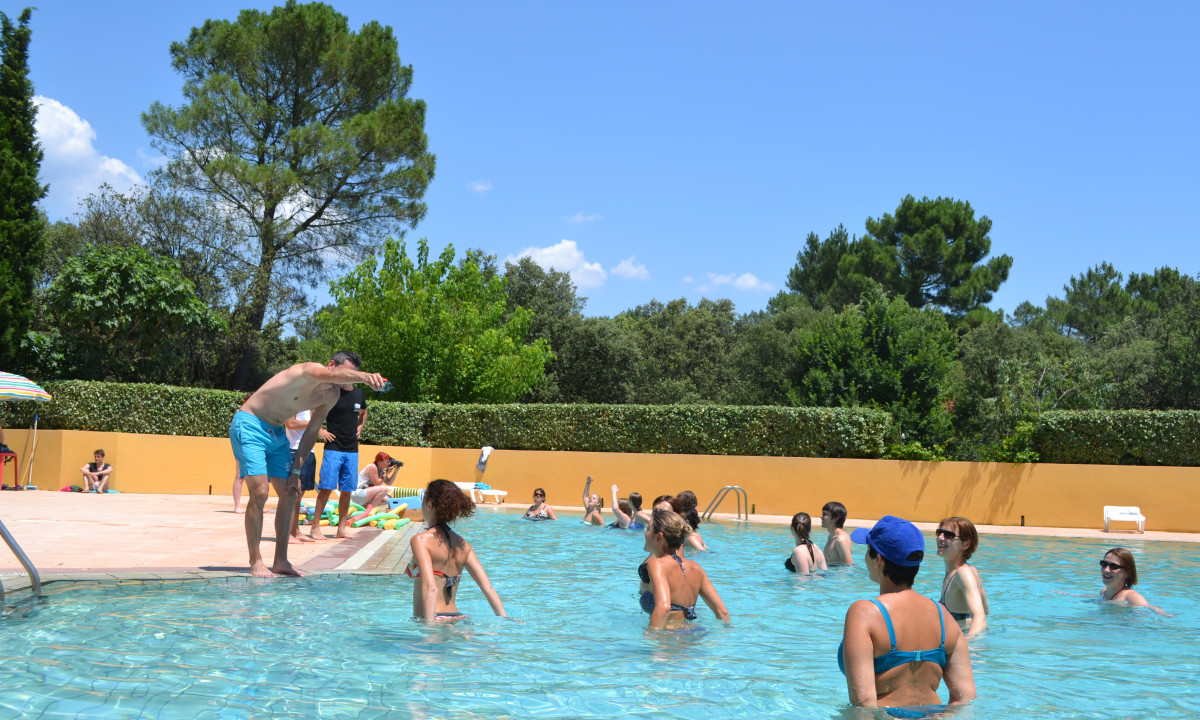 At La Sousta, individuals are swimming and unwinding in an outdoor pool, encircled by lush greenery beneath a clear blue sky.
