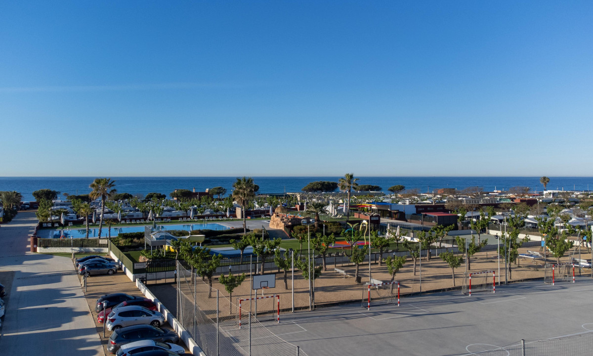 Basketball and soccer courts sit unused; poolside lounge chairs face the ocean under a clear blue sky at Resort Els Pins