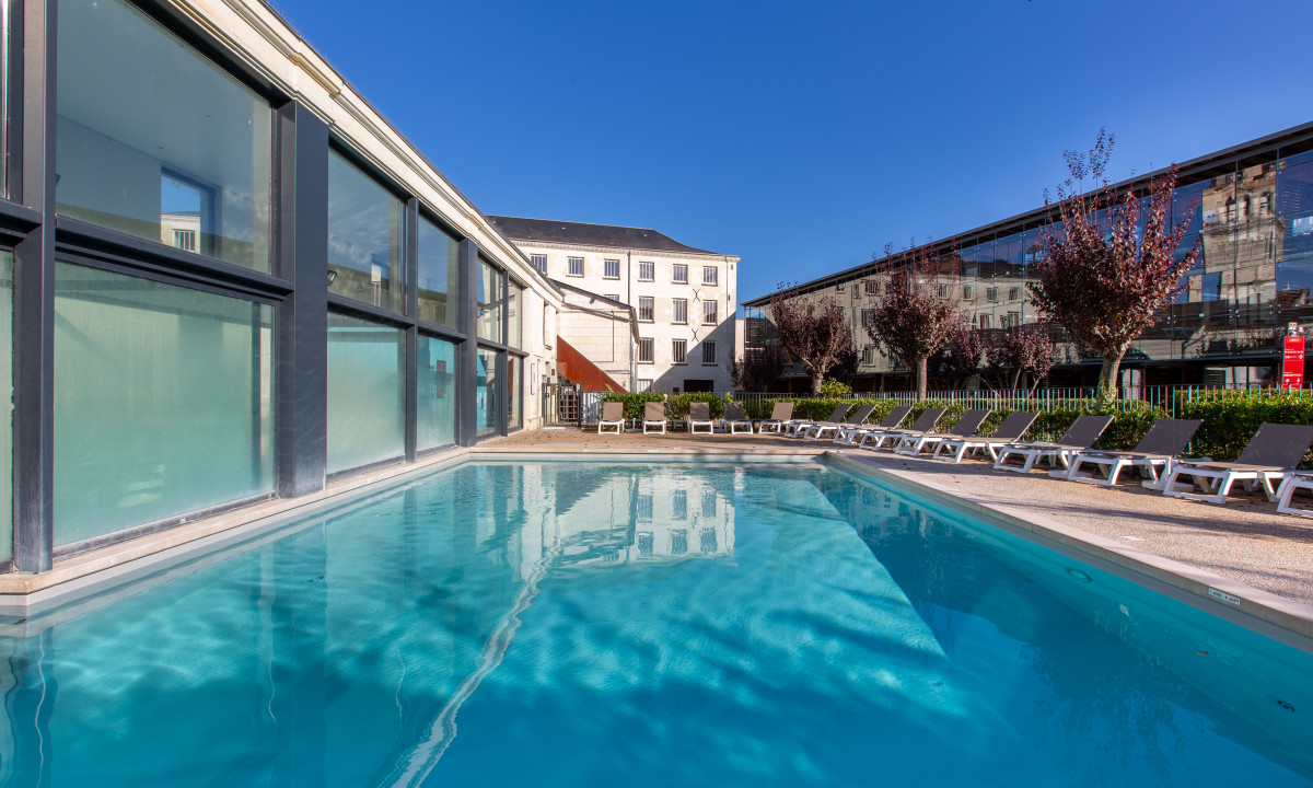 Swimming pool surrounded by sun loungers and modern buildings under a clear blue sky at Le Moulin des Cordeliers