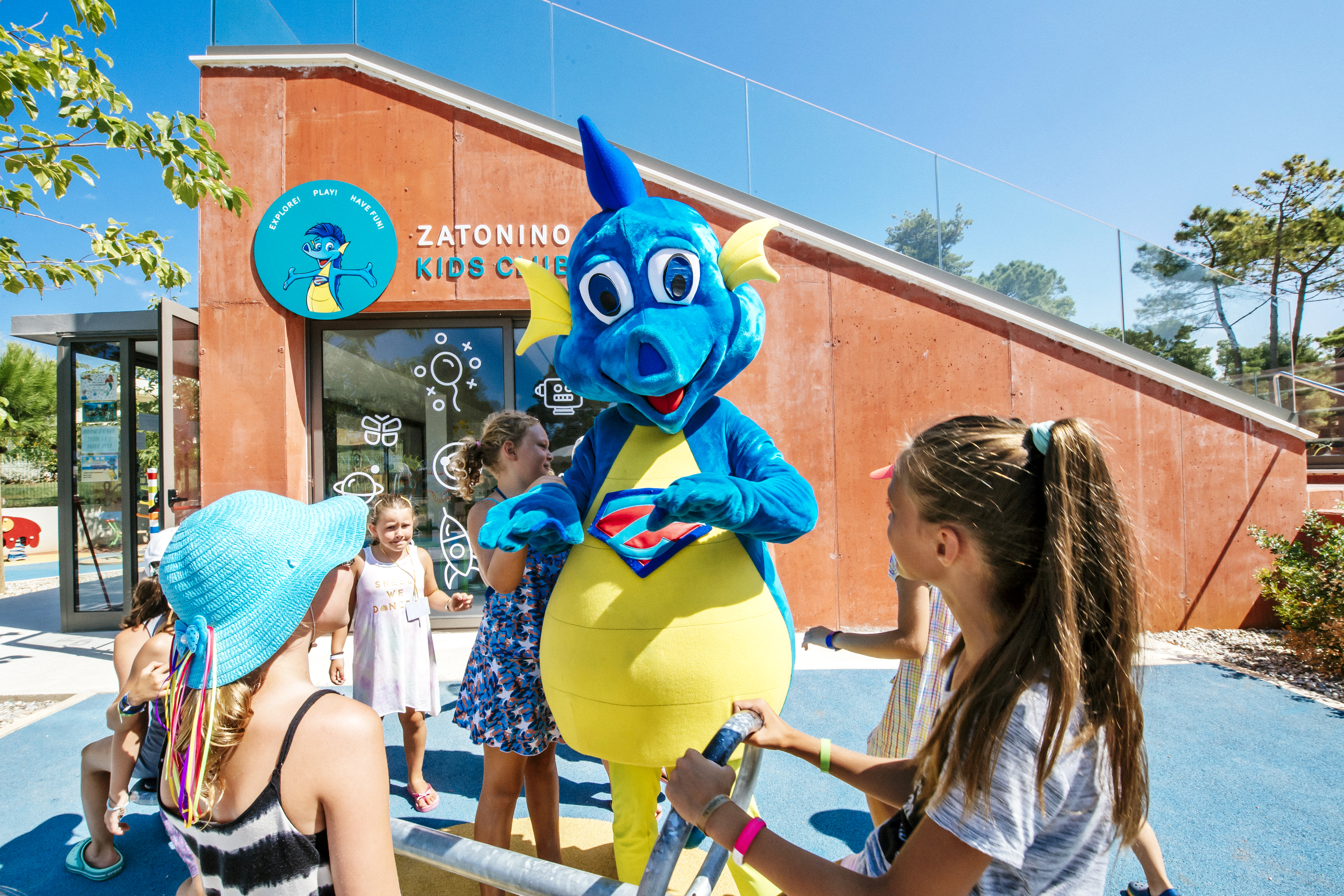 At Zaton Holiday Resort, a cheerful blue dragon mascot engages playfully with children outside a vibrant building on a sunny day.