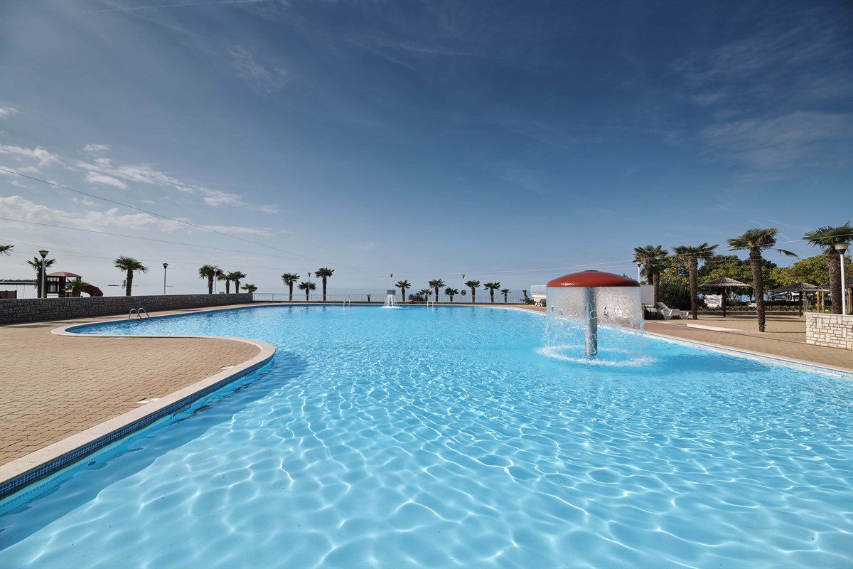 At Naturist Ulika, a spacious swimming pool with a water feature is encircled by palm trees and sun loungers, providing a view of the ocean.