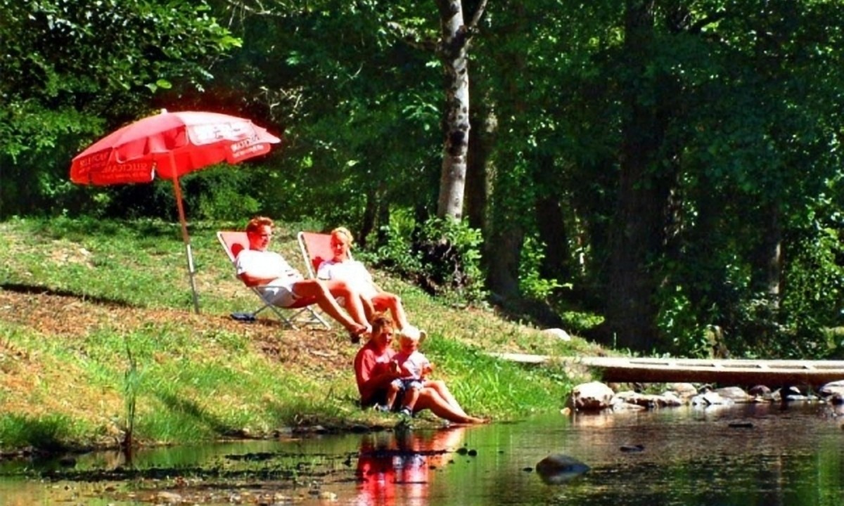 At Les Ranchisses, individuals are unwinding beneath a crimson umbrella, seated beside a woodland stream.