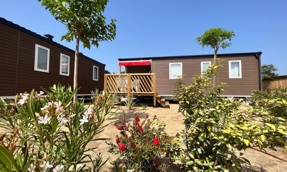 Cabin with porch surrounded by flowering bushes and small trees, set under a clear blue sky at Resort Els Pins