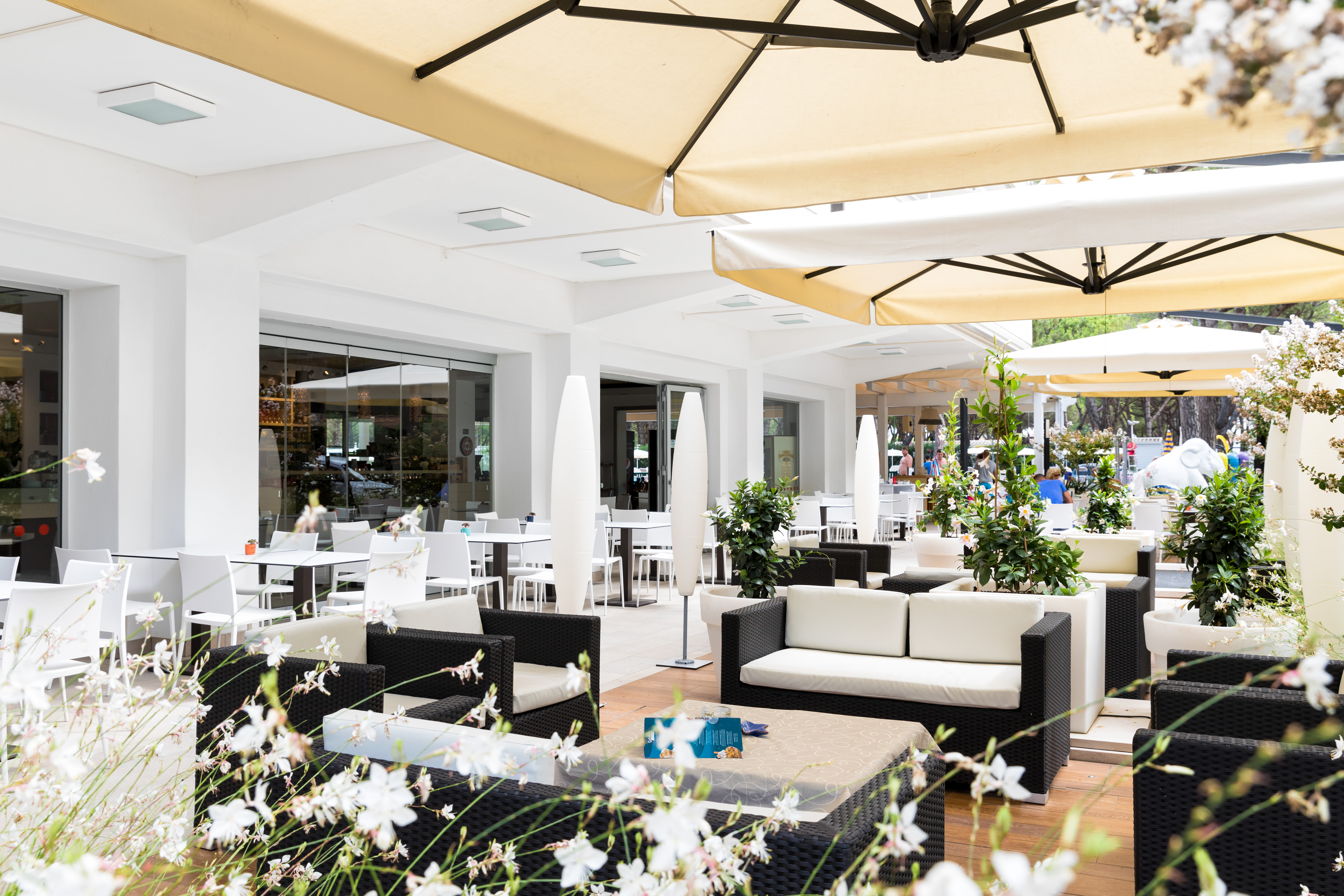 Outdoor lounge area featuring cushioned wicker seating and tables beneath large beige umbrellas amid white flowers and greenery at Residence Village