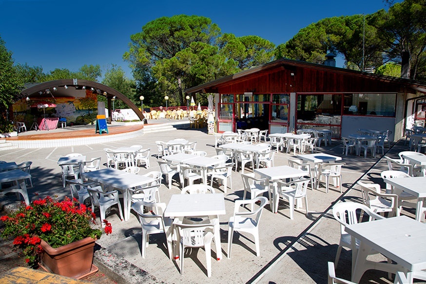 White tables and chairs are arranged outside a wooden building near a stage with trees in the background at Belvedere Pineta