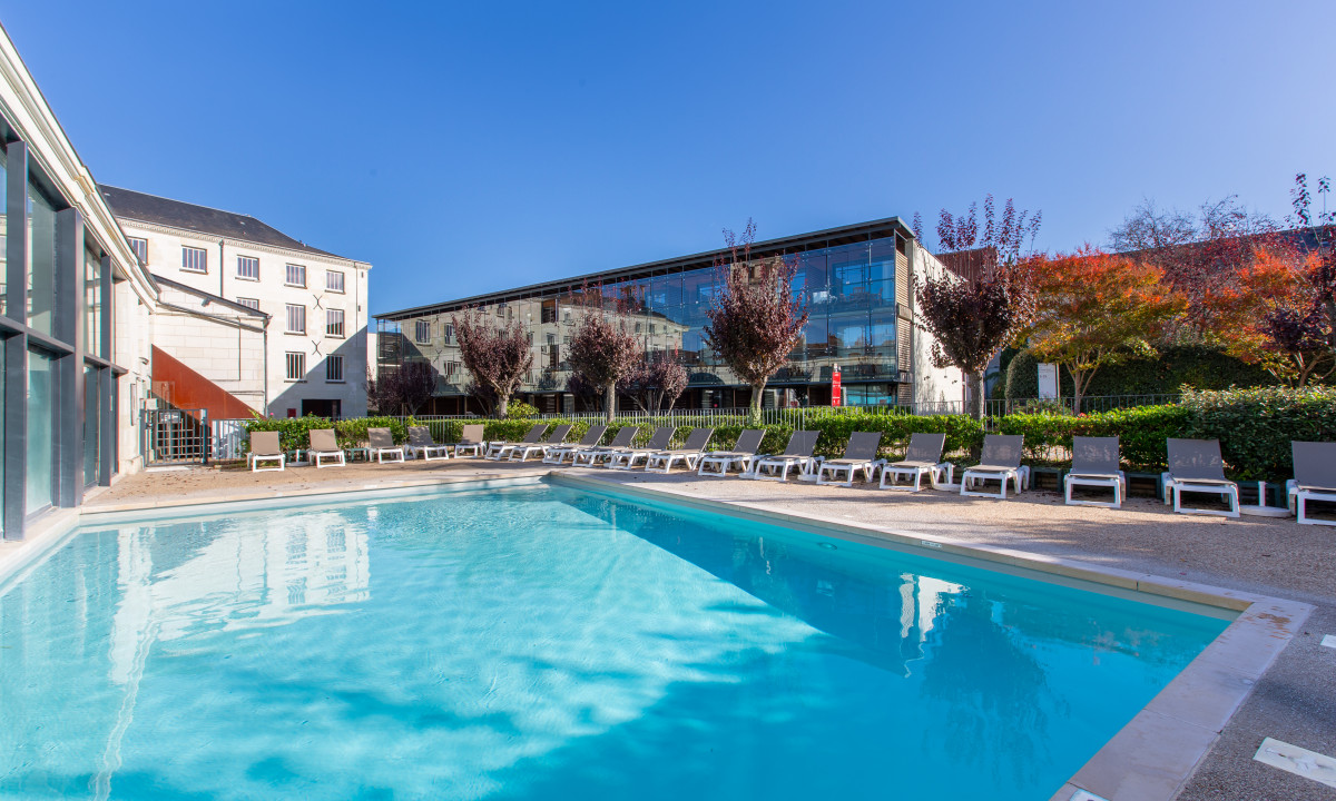 An empty swimming pool with surrounding lounge chairs in front of modern buildings and trees at Le Moulin des Cordeliers