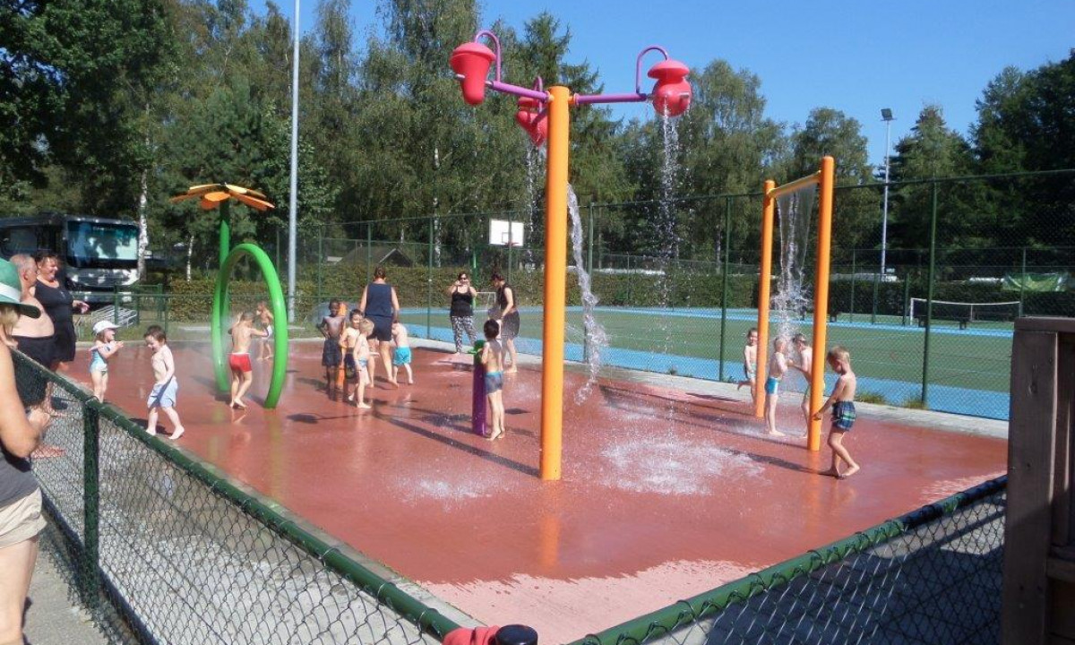 At het Veen, youngsters are enjoying themselves at a water splash pad, overseen by adults, amidst trees and a tennis court.
