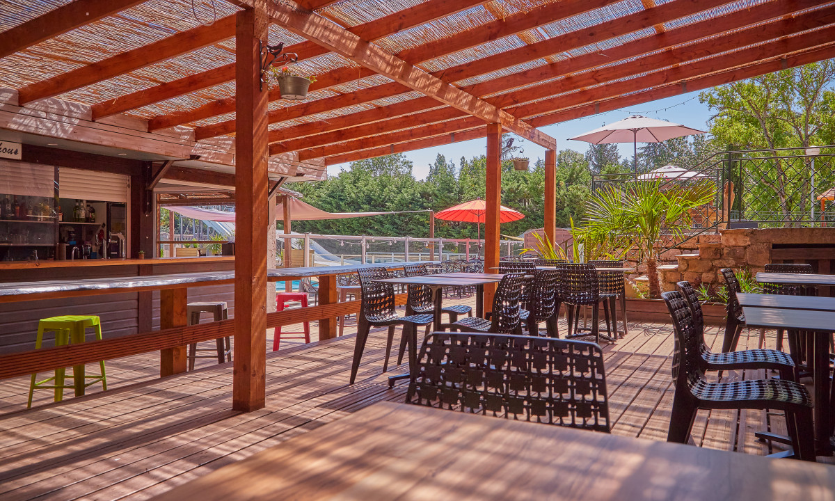 At Le Chêne Vert, there is an outdoor dining space shaded by a wooden pergola, showcasing black chairs and tables with lush green trees in the backdrop.