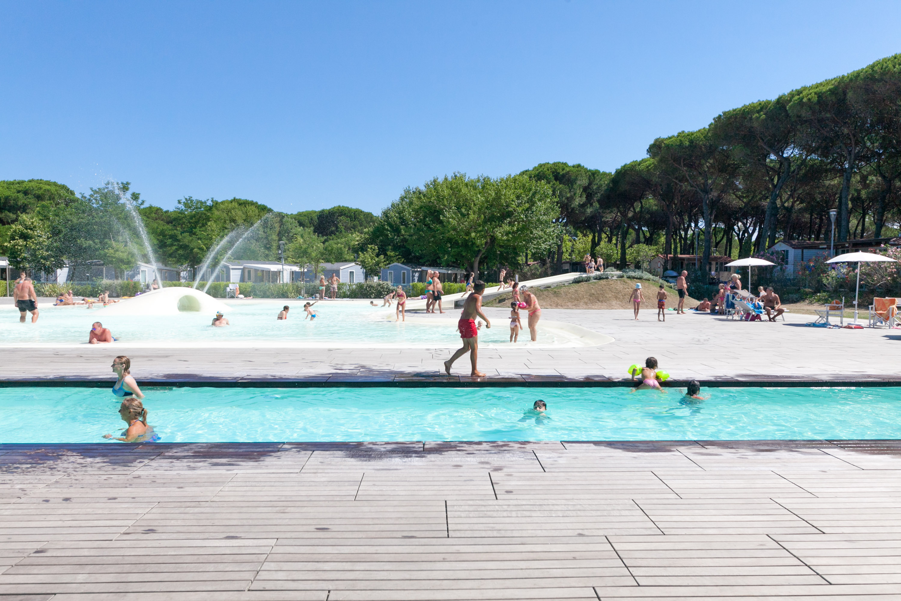 People swimming and playing in a large pool area surrounded by lush trees and cabins at Pineta Sul Mare Camping Village