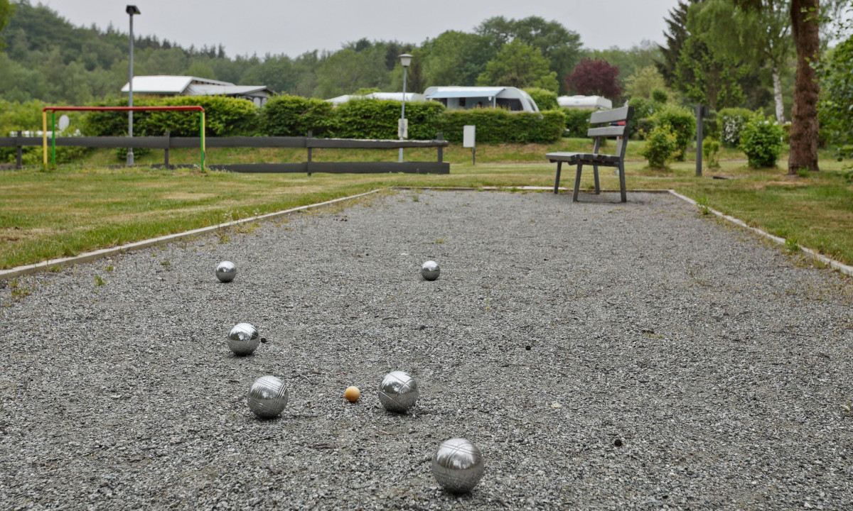 At KNAUS Campingpark Wingst, balls are dispersed on a gravel court, close to wooden seating and lush vegetation.
