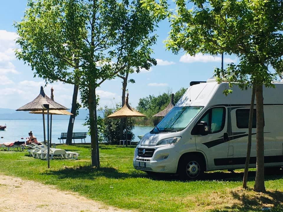 Campervan parked beside a lake, surrounded by trees and straw umbrellas; people relax by the shore at Punta Navaccia