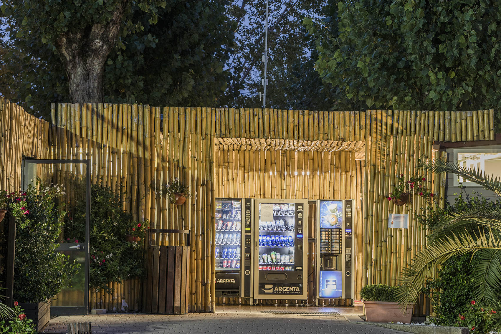 Vending machines dispensing snacks and drinks, enclosed by a lighted bamboo structure, surrounded by plants at hu Venezia Camping in Town