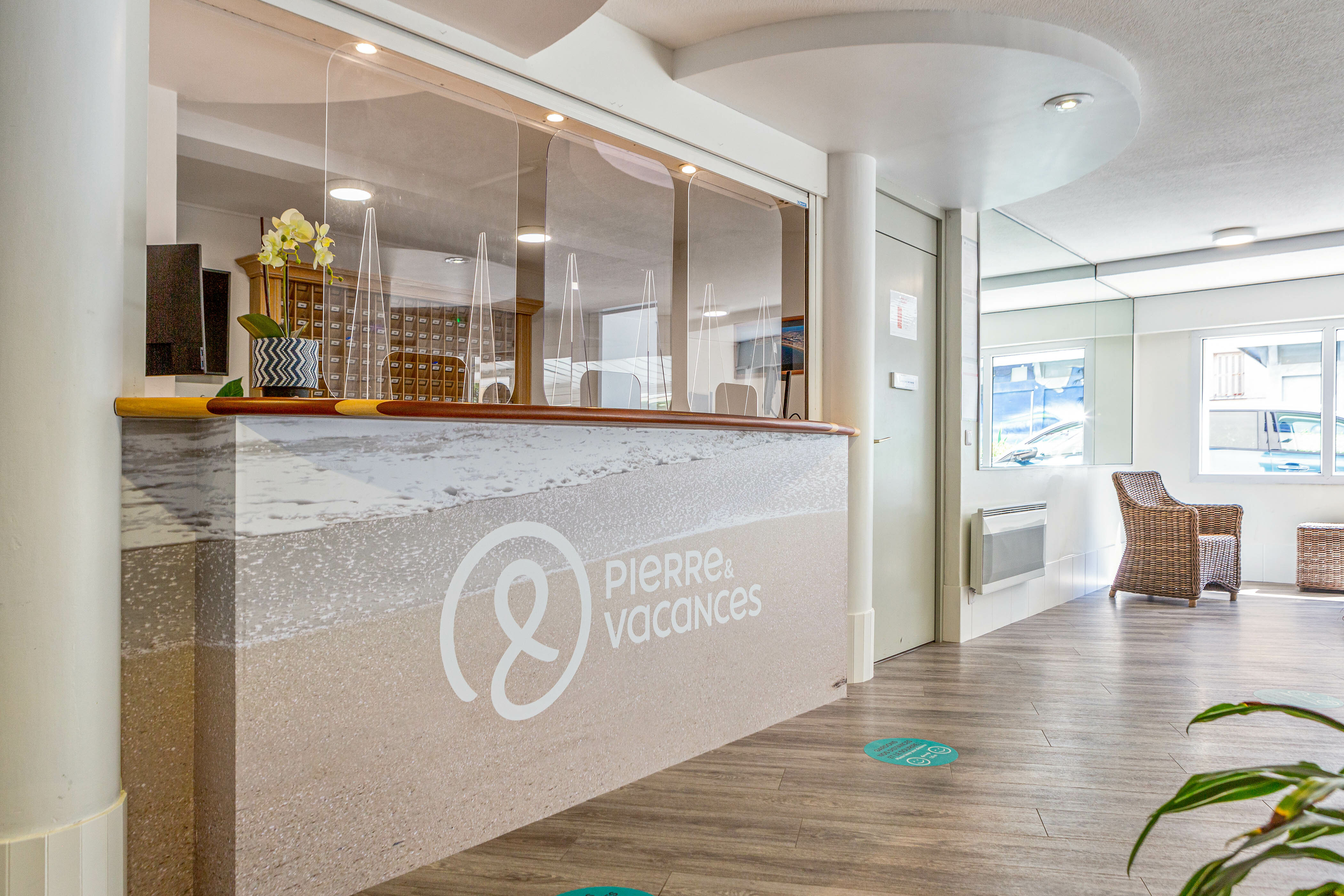 Reception desk with protective screens and a potted plant in a bright, modern lobby at La Baie des Sables
