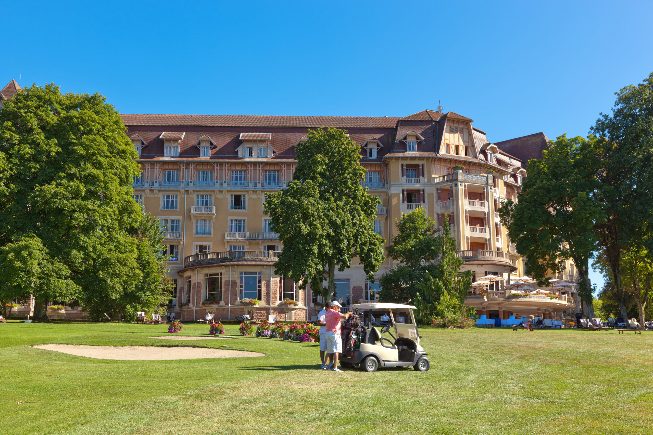 Golf cart parked on green lawn near large historic building with trees, bushes, and people in background at Seasonova Vittel