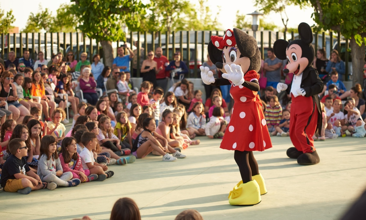 Two costumed characters dancing on stage, surrounded by seated children and adults, at a campsite event at Resort Els Pins