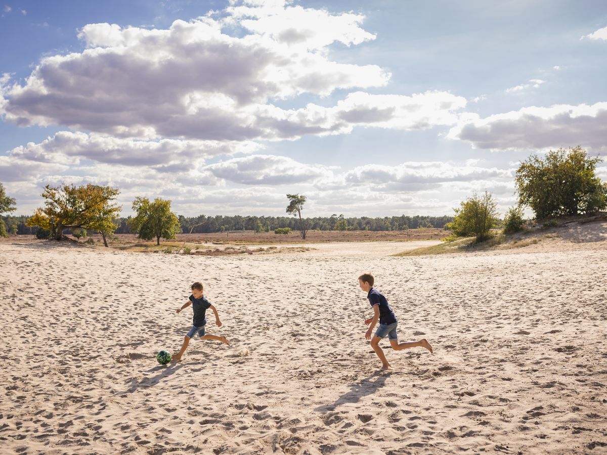 Two children run on sandy terrain, kicking a ball under a partly cloudy sky, surrounded by sparse trees at Marvilla Parks Kaatsheuvel