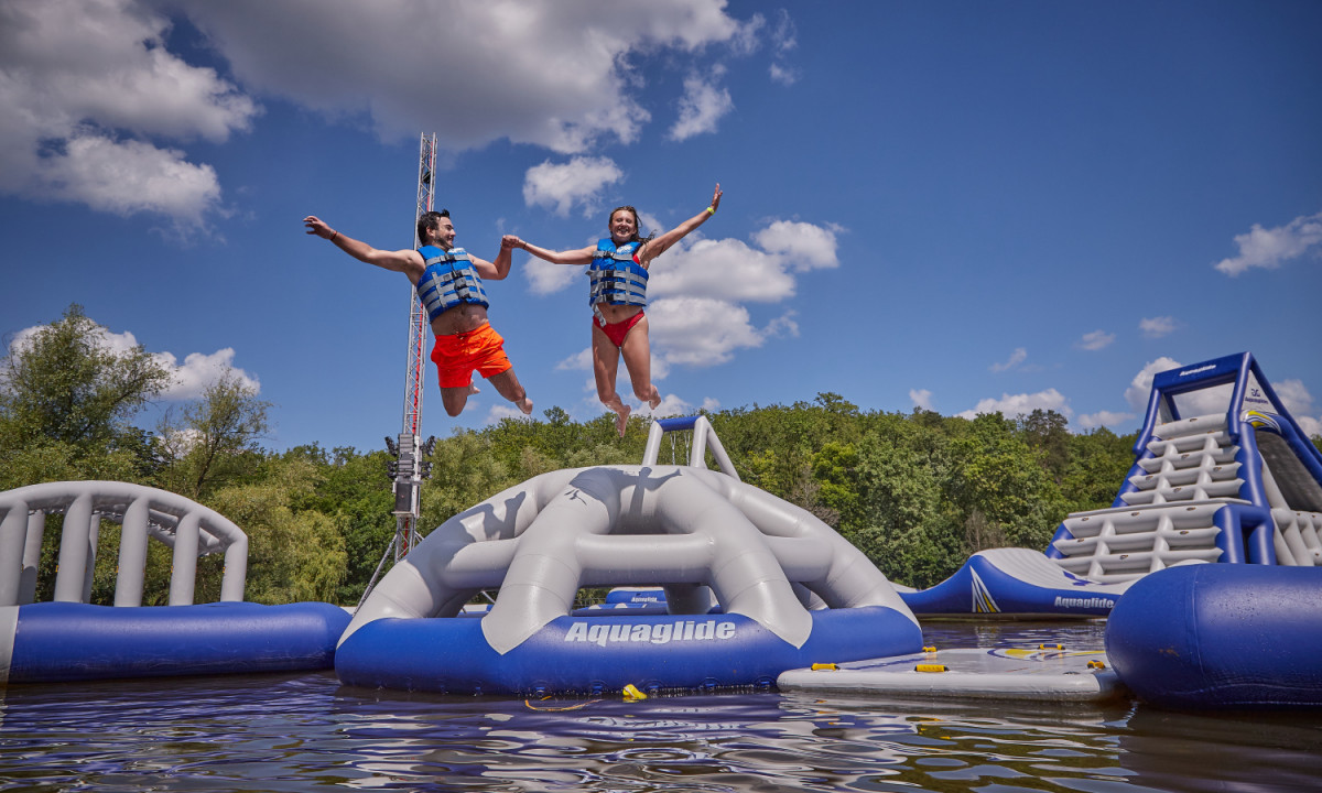 At Coucou, two individuals leap from an inflatable aquatic structure, donning life vests, encircled by trees and a clear azure sky.