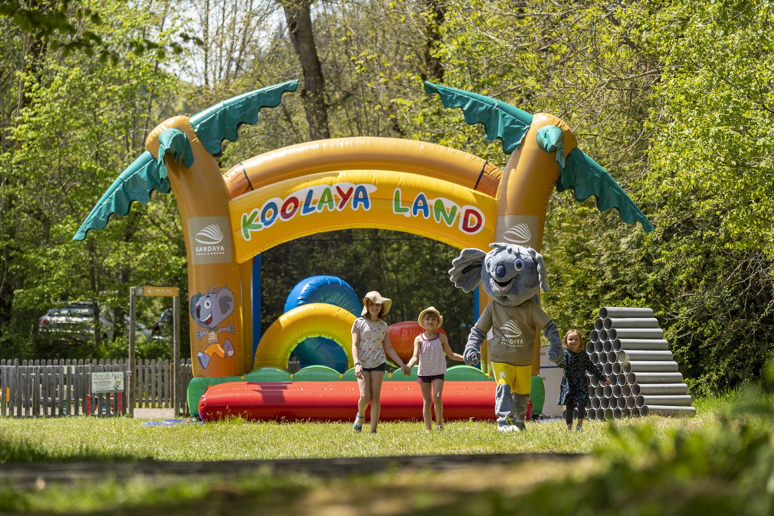 At La Ribeyre, an inflatable archway is present with children and a mascot strolling through a grassy space adjacent to a forest.