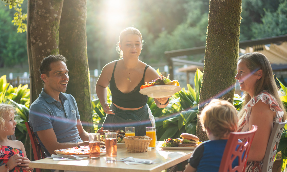 At La Clémentine, an individual presents meals to a family dining al fresco, enveloped by vibrant foliage and sunlight.