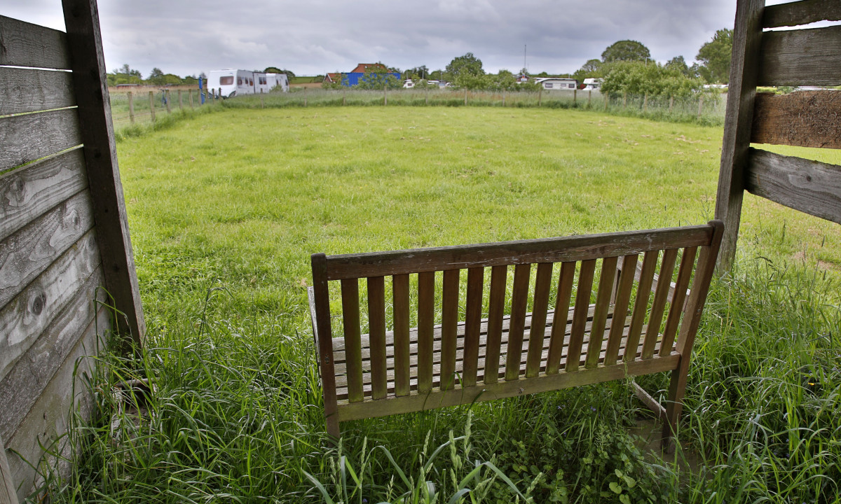 At KNAUS Campingpark Eckwarderhörne, a timber bench rests on the grass inside a wooden pavilion, oriented towards a verdant field with caravans in the distance.