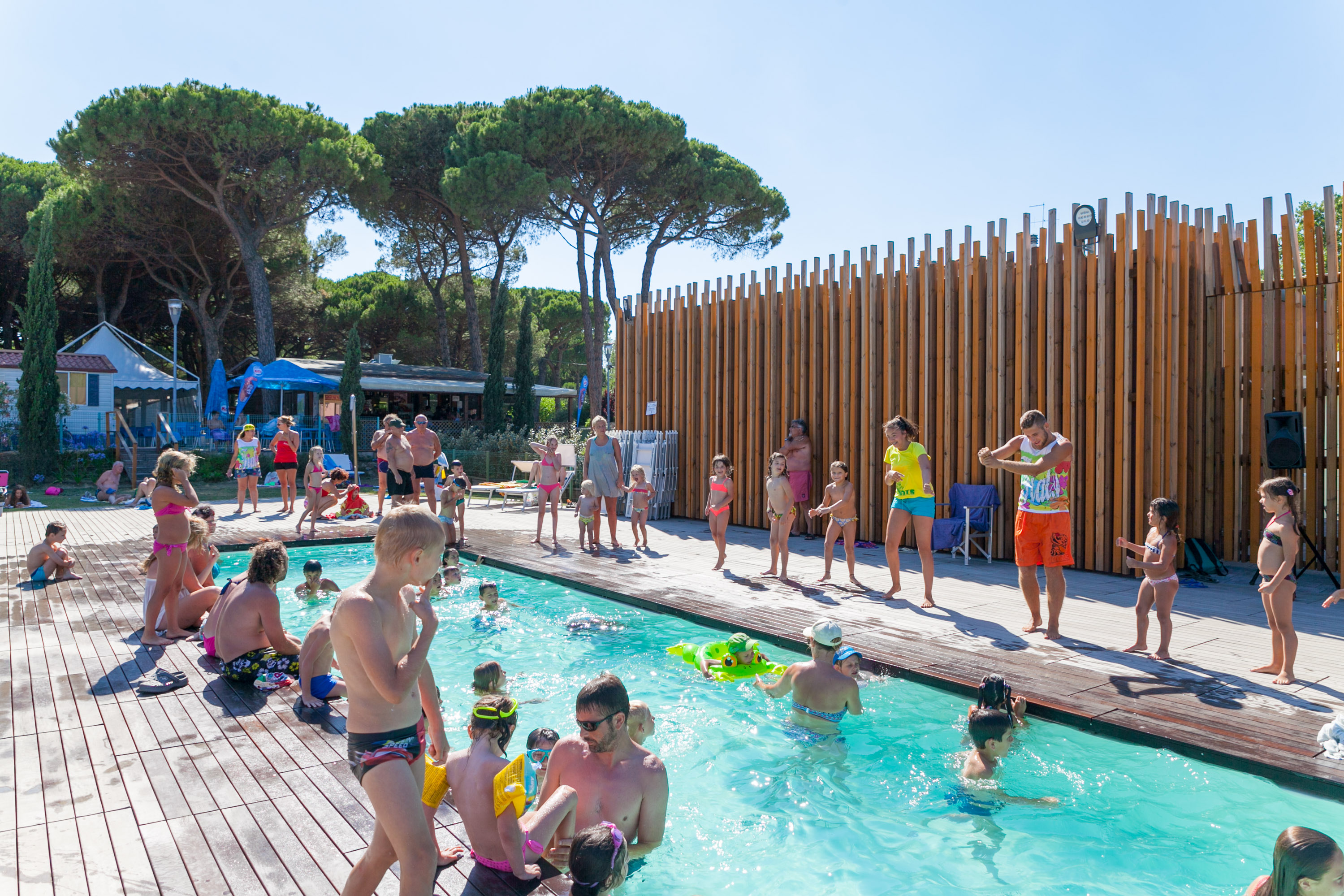 People swimming and enjoying a pool while others participate in a dance activity by a wooded area at Pineta Sul Mare Camping Village