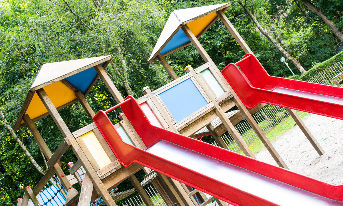 At het Veen, there is a wooden play structure featuring red slides and vibrant roofs situated in a wooded area.