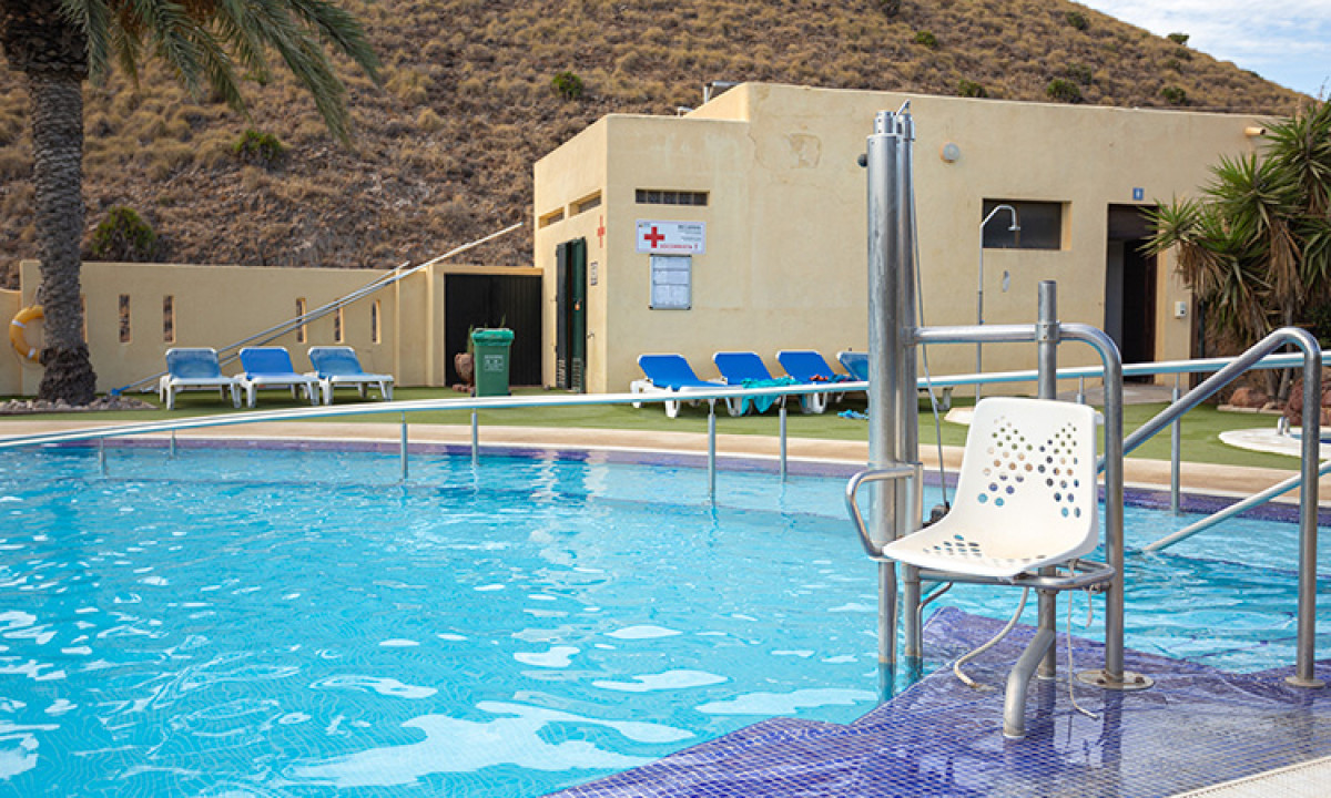 At Camping Los Escullos, there is a poolside lift chair next to the swimming pool, with lounge chairs and a structure in the background.