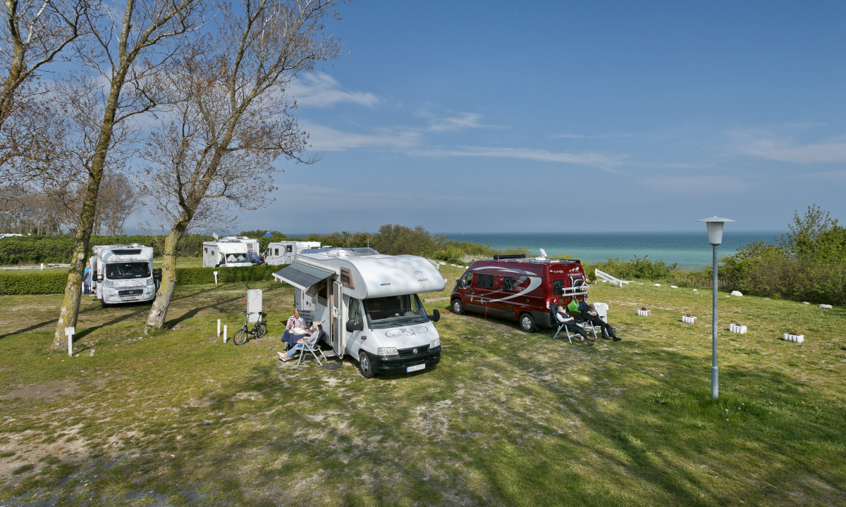 At KNAUS Campingpark Rügen, campers lounge outside their RVs, unwinding beneath trees close to the sea, encircled by grassy spaces adorned with scattered lampposts.