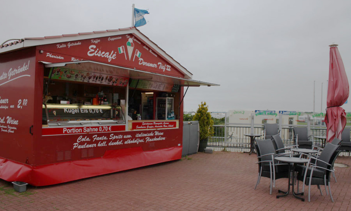 At KNAUS Campingpark Dorum, a food truck offers ice cream and beverages, encircled by patio seating and collapsed umbrellas in a cloudy outdoor environment.