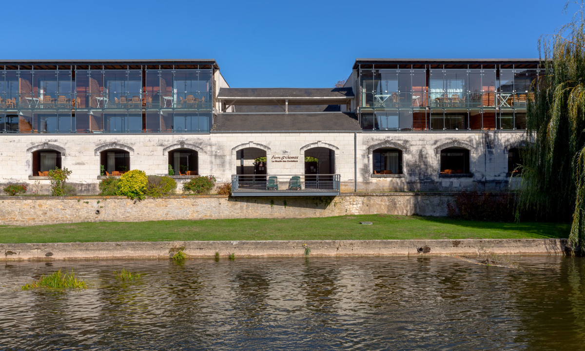 Building with large windows overlooking a river, surrounded by greenery and flowers at Le Moulin des Cordeliers