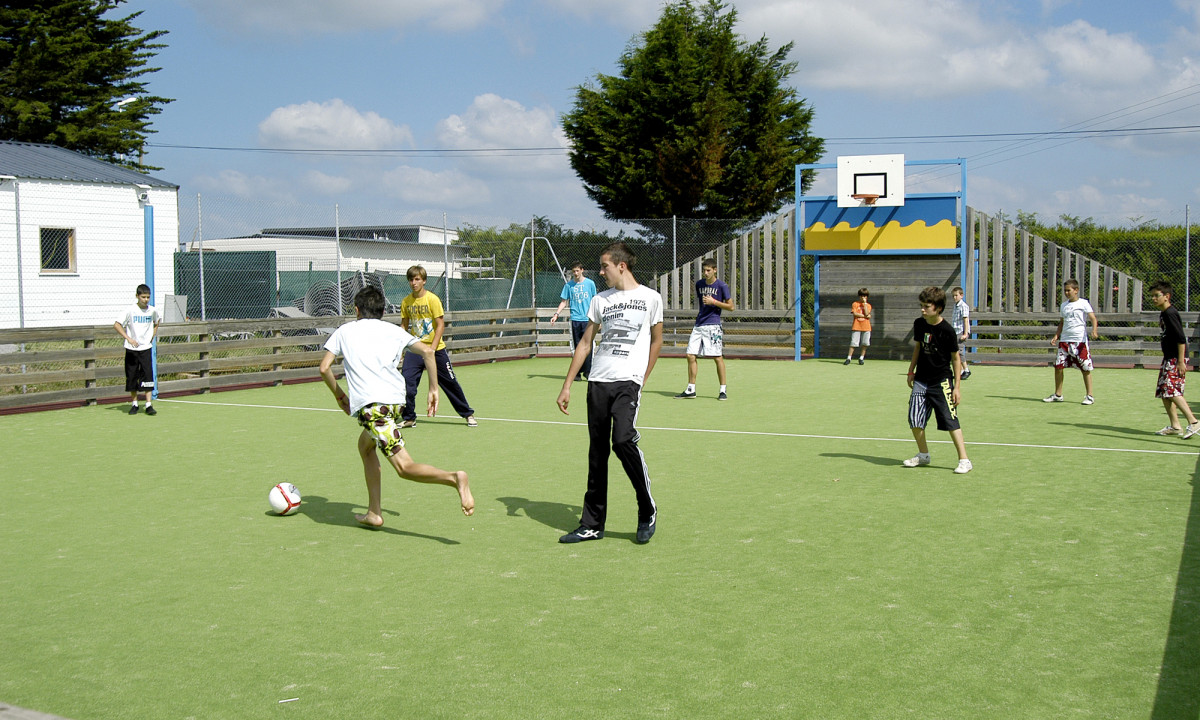 At Le Domaine de Léveno, kids are playing soccer on an outdoor pitch enclosed by a wooden barrier, with a basketball hoop nearby.