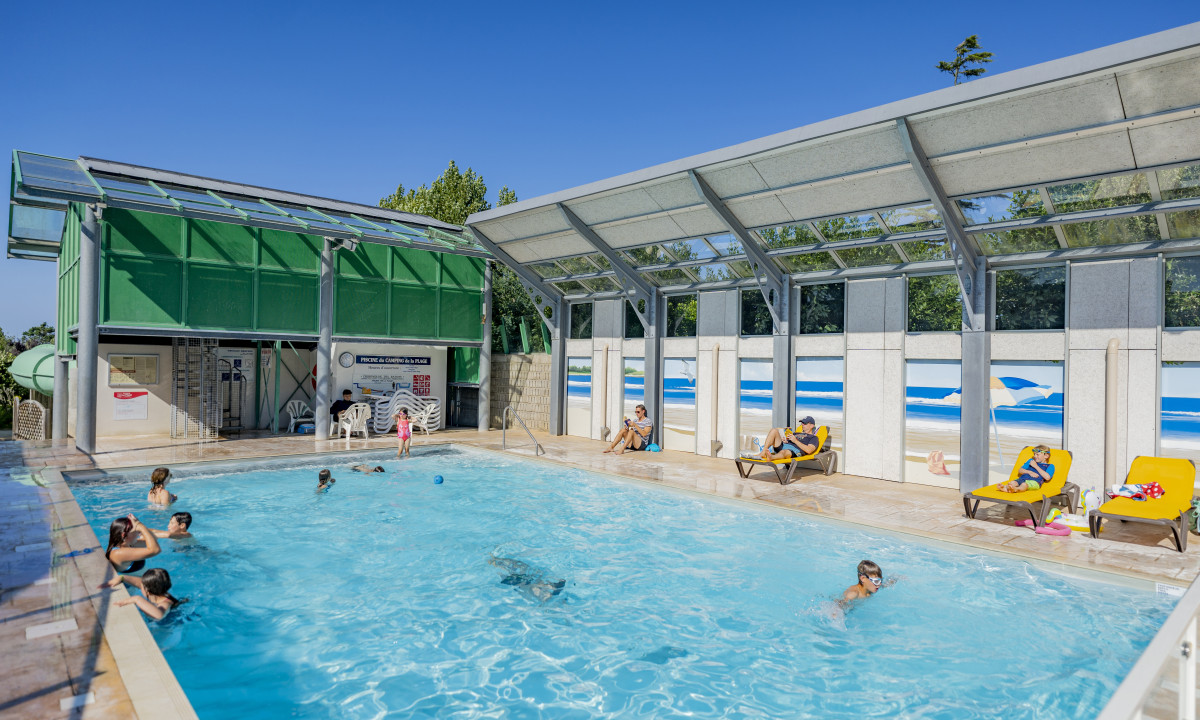At De la Plage, an indoor swimming pool features individuals swimming and unwinding on lounge chairs, beneath a glass ceiling, adorned with beach-themed murals.