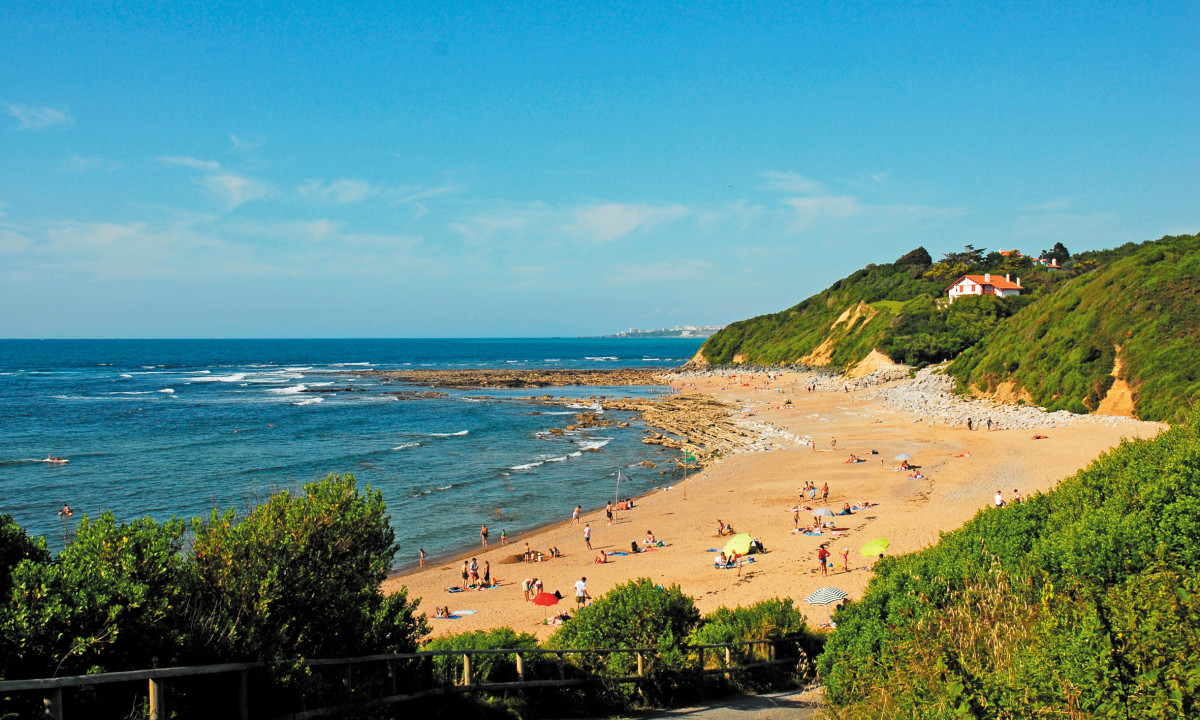 Beachgoers sunbathing and swimming on a sandy beach next to green hills and a house at Atlantica