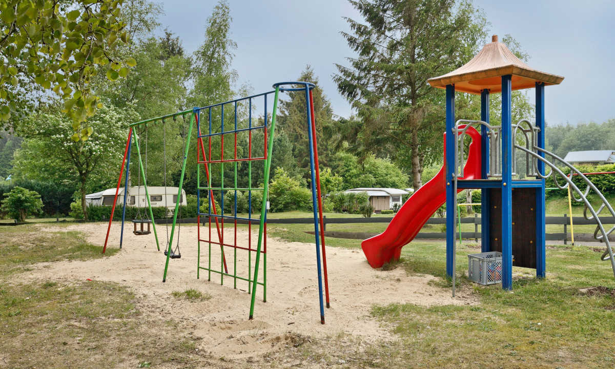 At KNAUS Campingpark Wingst, there is a play area featuring swings and a slide situated in a sandy space, encircled by trees and caravans.