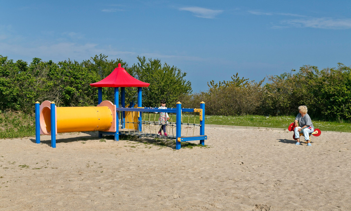 At KNAUS Campingpark Rügen, vibrant play structures feature a slide, a child traversing a rope bridge, and an adult sitting on a spring rider, all amidst lush greenery.