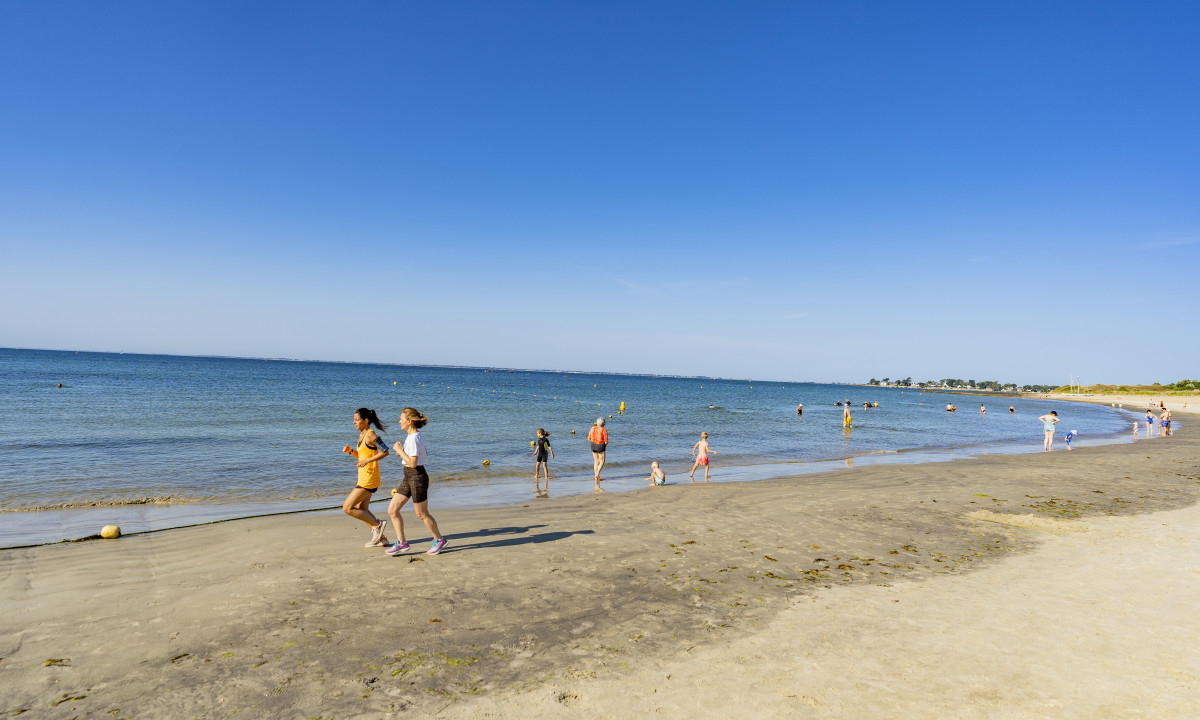 At De la Plage, two individuals run along a sandy shoreline, while swimmers and sunbathers relish a bright day by the ocean.