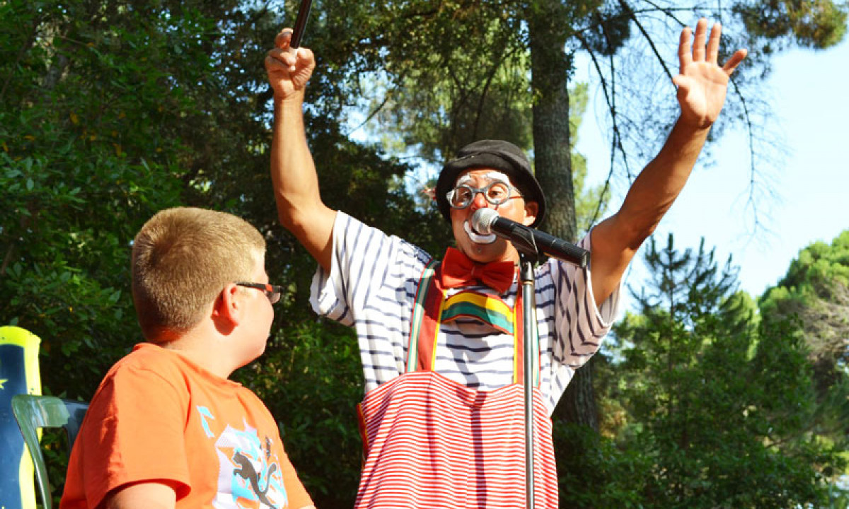 At La Sousta, a clown amuses a seated child with lifted arms and props surrounded by towering trees and lush greenery.