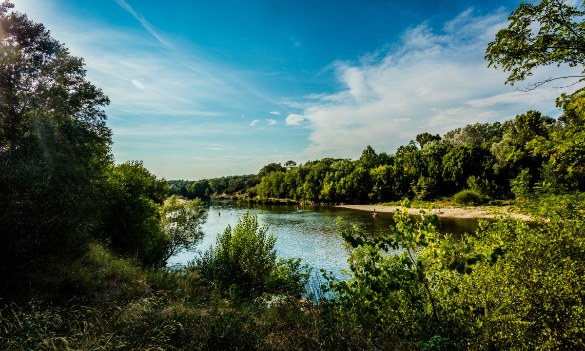At La Sousta, a river mirrors the sky, bordered by vibrant trees and shrubs, with individuals along the riverbank.
