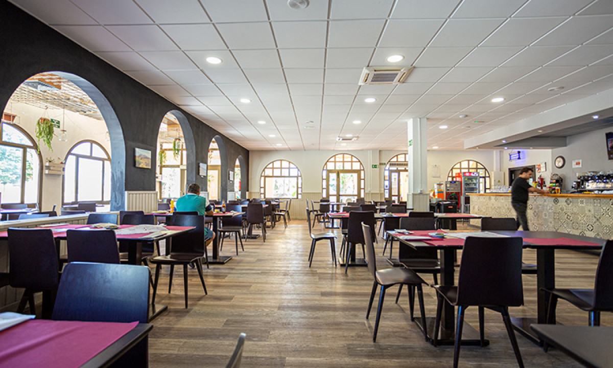 At Camping Los Escullos, the restaurant's interior features tables and chairs organized in rows, a person strolling near the counter, and large arched windows.
