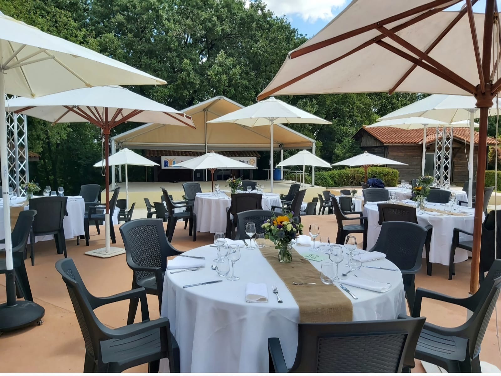 At Domaine du Koukano, tables shaded by umbrellas are arranged with tableware in an outdoor dining space, encircled by lush greenery and buildings.